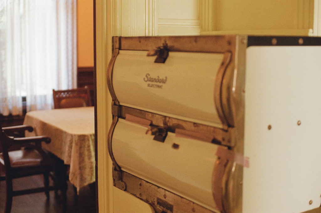 Vintage white Standard Electric stove with curved doors in the foreground, dining room with wooden table and chairs visible through a doorway in the background
